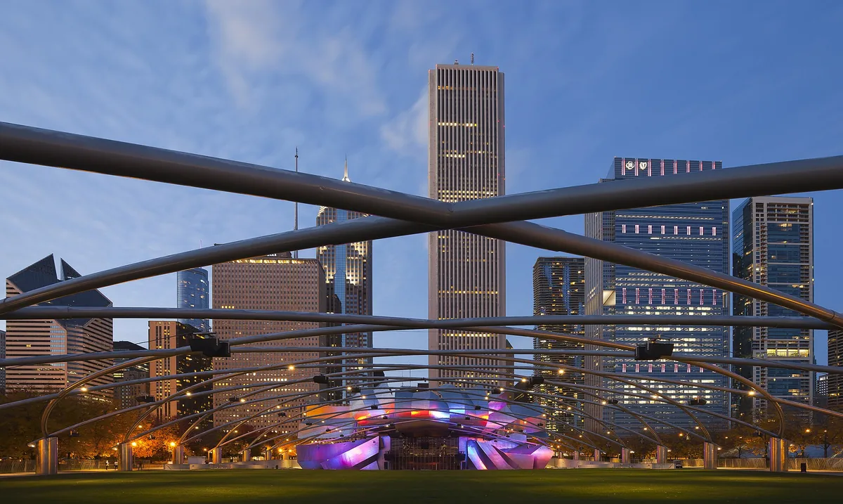 The steel trellis of the Jay Pritzker Pavilion in Millennium Park at dusk, with the downtown skyline behind it