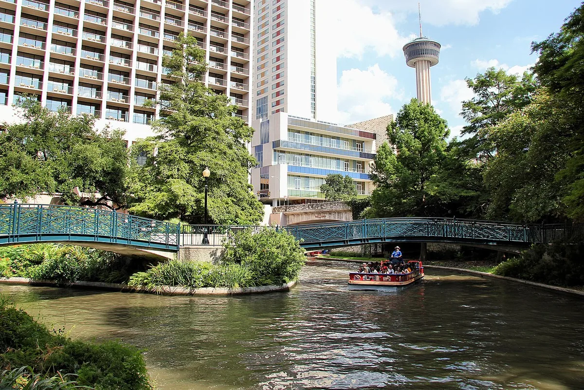 A river barge passing under a footbridge on the San Antonio River Walk with the Tower of the Americas behind